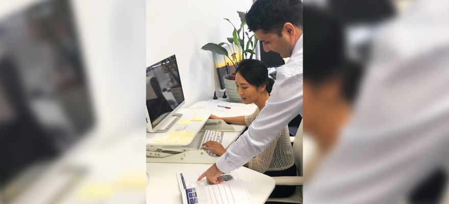 A woman is working at a computer while her colleague points to a printed graph on the table beside her