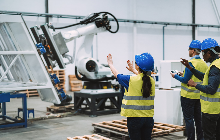 A large robotic arm is lifting a large panel, while three people in safety helmets and yellow vests stand facing it nearby with their backs to the camera.