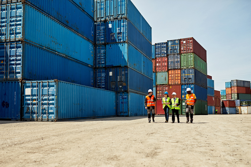 From a distance we see four people with yellow and orange vests walking together through a shipping container storage area.
