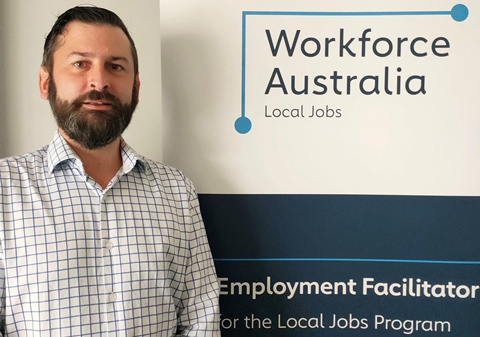 Luke Arber, the Employment Facilitator for Gippsland, stands wearing a business shirt in-front of a Workforce Australia banner.