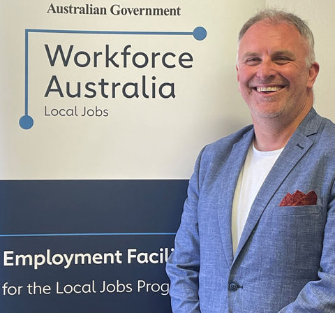 The Employment Facilitator for Hunter, Grahame Irwin, stands smiling in a blue suit jacket in-front of a Workforce Australia banner.