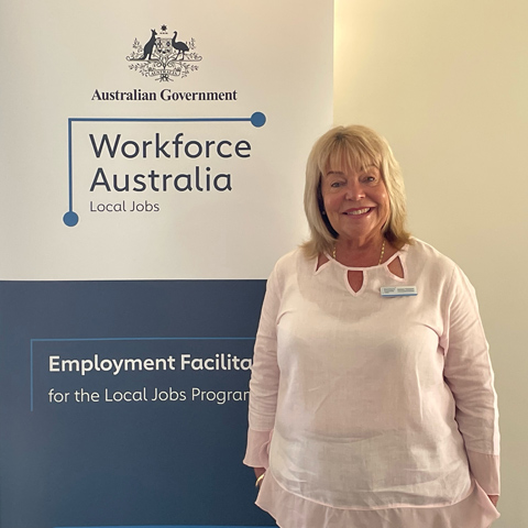 The Employment Facilitator of Geraldton, Barbara Thompson, faces the camera smiling as she stands in front of a Workforce Australia banner.