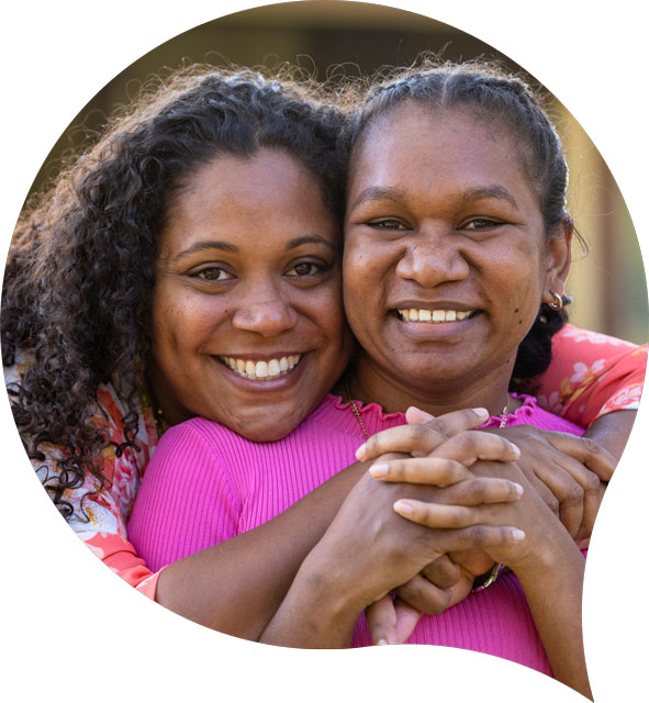 Mid-close up of two women smiling at the camera as they embrace.