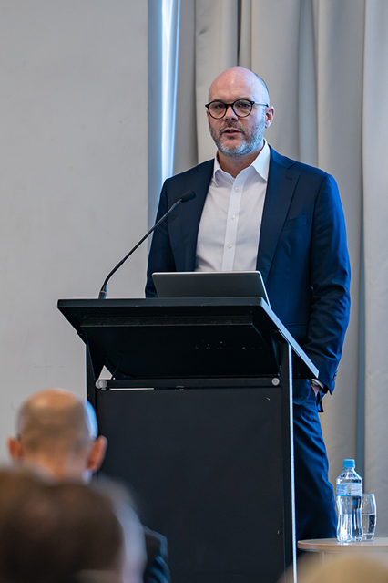 Matt Cowgill stands behind a podium, delivering a presentation to a room of people with a projector screen showing a data slide behind him.