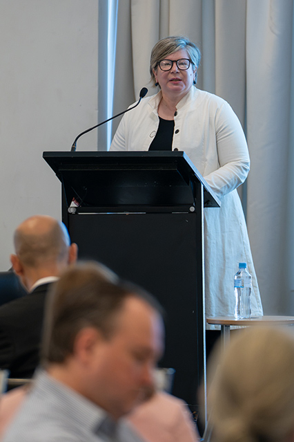 Tania Rishniw stands behind a podium, delivering a presentation to a room of people with a projector screen showing a data slide behind him.