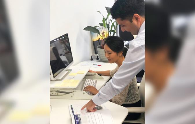 A woman is working at a computer while her colleague points to a printed graph on the table beside her