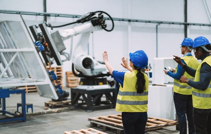 A large robotic arm is lifting a large panel, while three people in safety helmets and yellow vests stand facing it nearby with their backs to the camera.