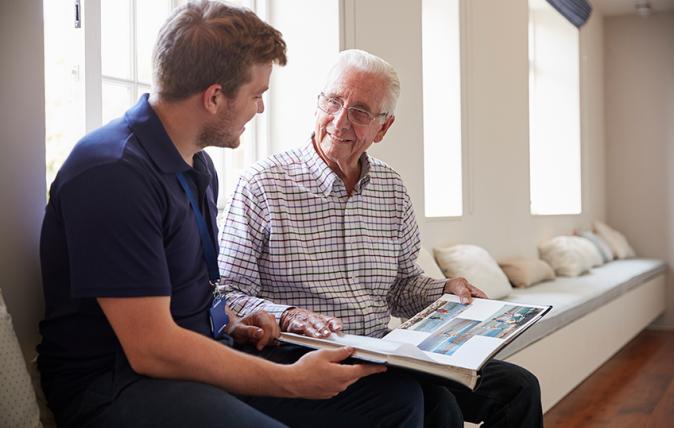 Mid-shot of an elderly gentleman seated next to a male nurse, to whom he is proudly showing a photo album.