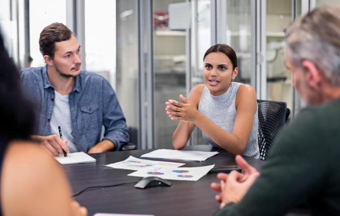 Young woman talking in an office business meeting while the three people around the table listen attentively.