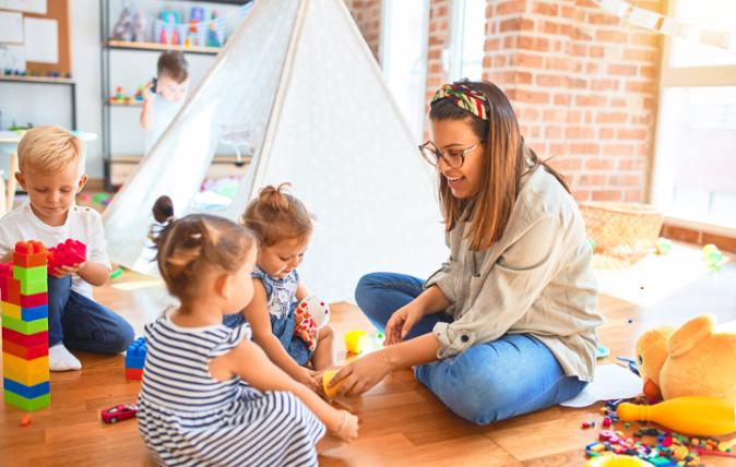 In a brightly lit room three young children are playing in front of their carer, a young woman who sits facing them