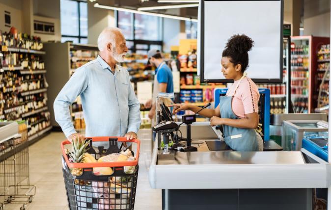 Employee in supermarket standing in front of a cash register serving senior customer