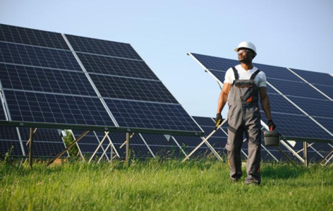 A woman wearing an orange high visibility vest with a matching hardhat is leaning on a solar panel, looking off-screen with a clear blue sky in the background.
