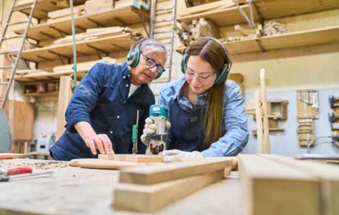 A woman and man in high visibility jackets and blue hardhats are in front of an open panel containing gauges and valves, discussing something about it.
