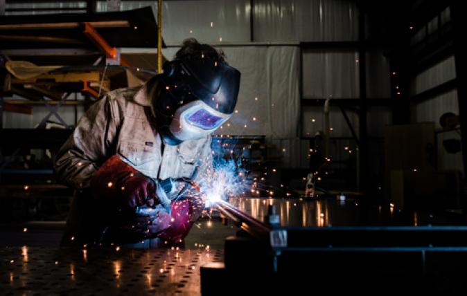 In a metal workshop, two young workers are looking down on the saw bench that one of them is working on.