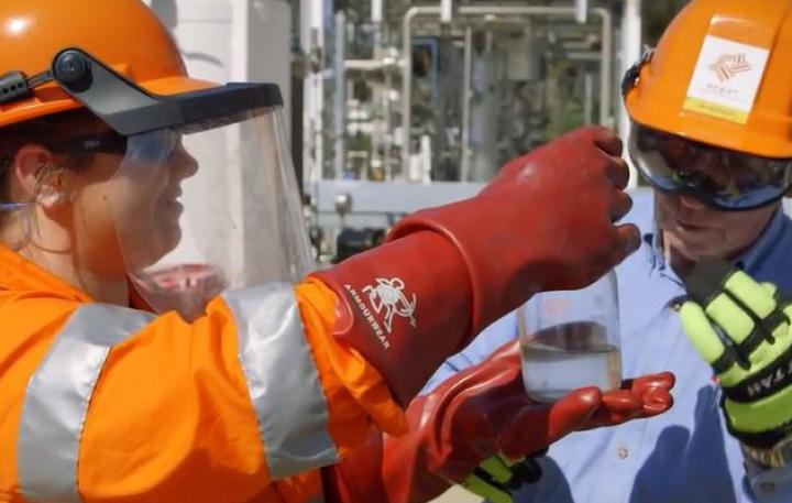 A student and her employer wearing safety helmets and standing in an outdoor construction environment examine a glass bottle containing a clear, liquid substance