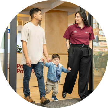 A woman wearing a maroon Parents Pathways shirt is walking out of a Parents Pathways centre, facing the man next to her who has his hand on the back of his young son between them.