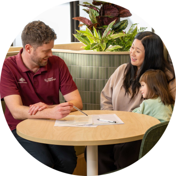 Around a small round table, a mother and young daughter sit next to a man wearing a maroon Parents Pathways shirt, who is holding a pen atop of printed forms.